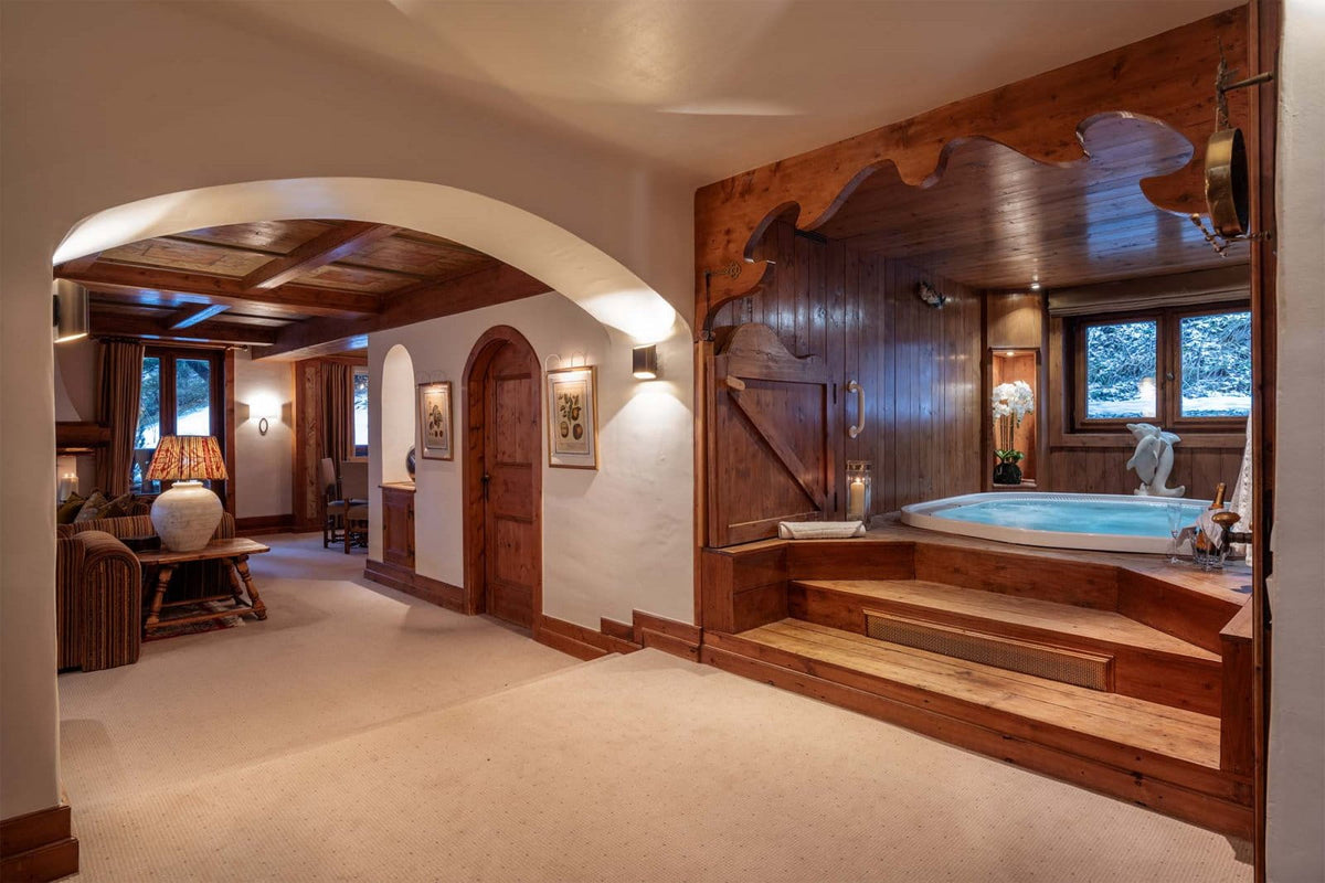 A luxury spa-style hot tub set in a rustic mountain chalet bathroom with warm wood paneling, stone walls, and an arched entryway leading to a living area.