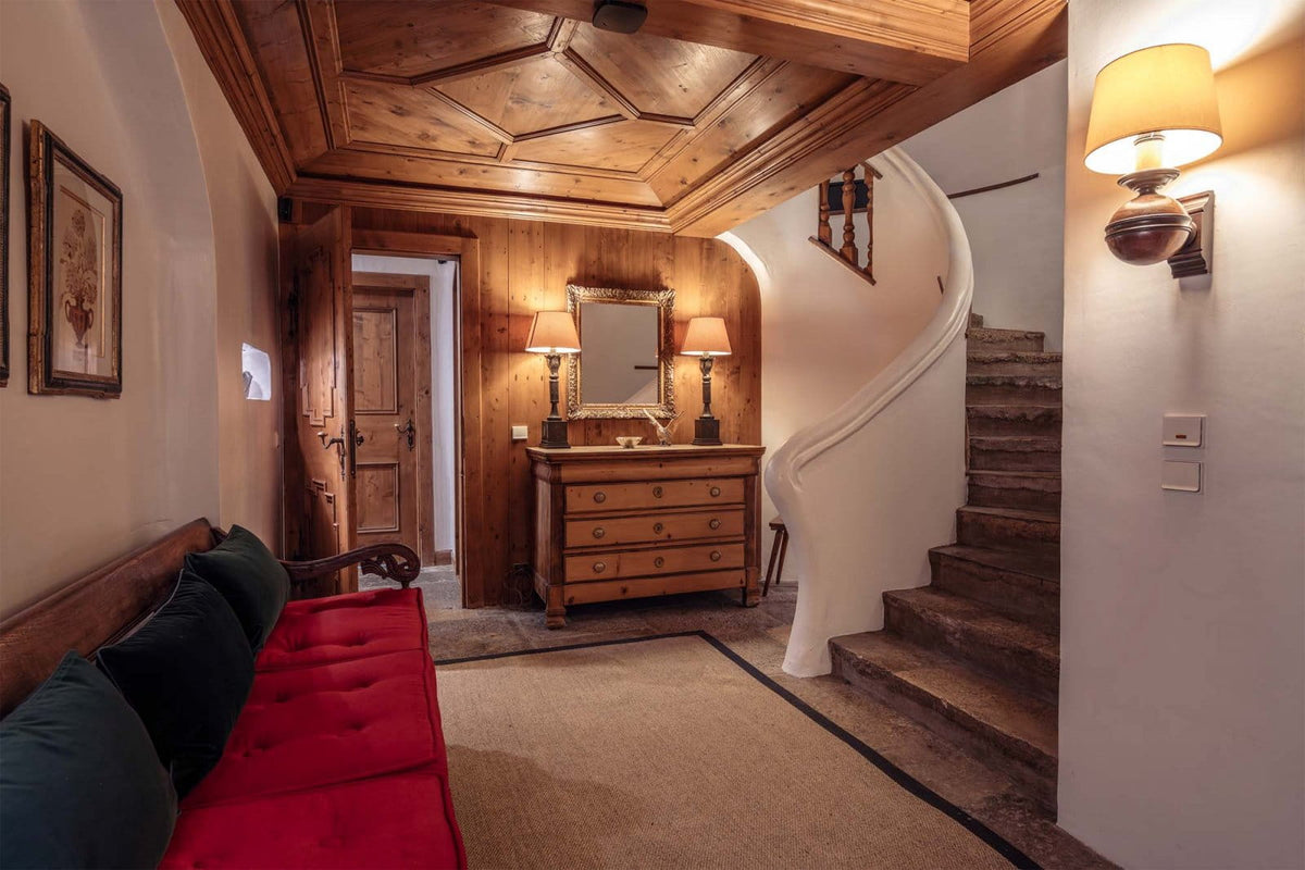 A cozy interior hallway with a curved white staircase, warm wood-paneled ceiling and walls, a wooden chest of drawers with lamps, framed wall art, and a red upholstered bench in the foreground.