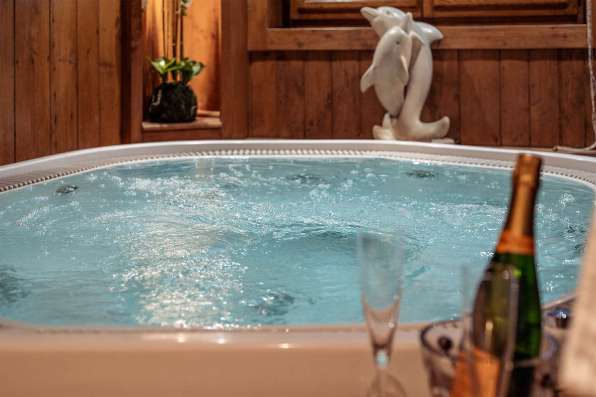 A bubbling hot tub with light blue water in a rustic wooden room, with a champagne bottle, glasses, and a white dolphin statue in the background.