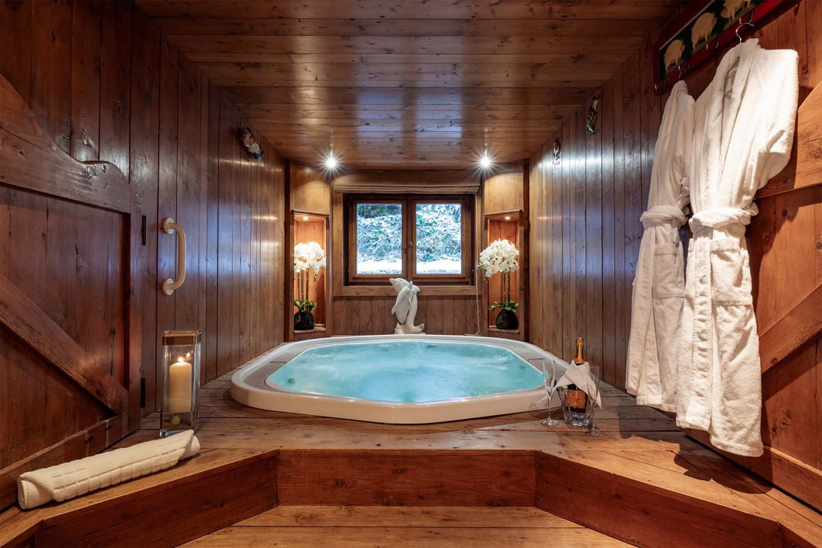 A rustic wooden chalet bathroom featuring a low, hexagonal hot tub with light blue water, white robes hanging on the right wall, and a window looking out onto a snowy forest.