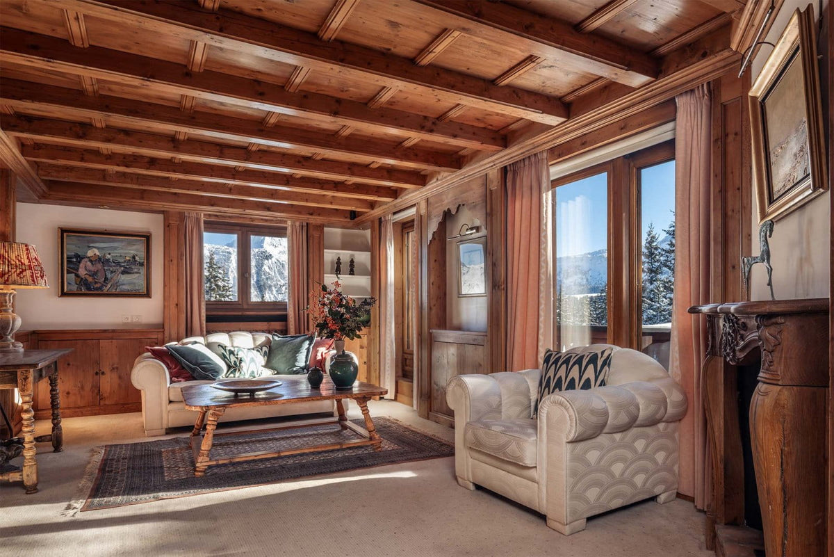 A luxurious ski chalet living room featuring a rustic wood-paneled ceiling, a cream tufted armchair, a matching sofa with decorative pillows, and large windows looking out onto snowy mountains.