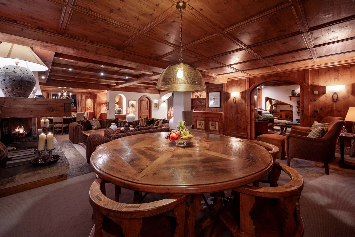 A rustic wooden dining room interior with a round wooden dining table and matching chairs in the foreground, a large dome pendant light hanging above, a fireplace on the left, and warm wood-paneled walls and ceiling in the background.