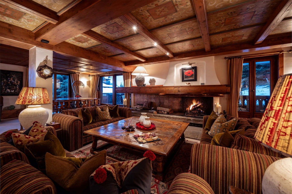 A cozy, rustic ski lodge living room with a large fireplace, wooden ceiling beams, striped armchairs, a wooden coffee table, and warm ambient lighting.