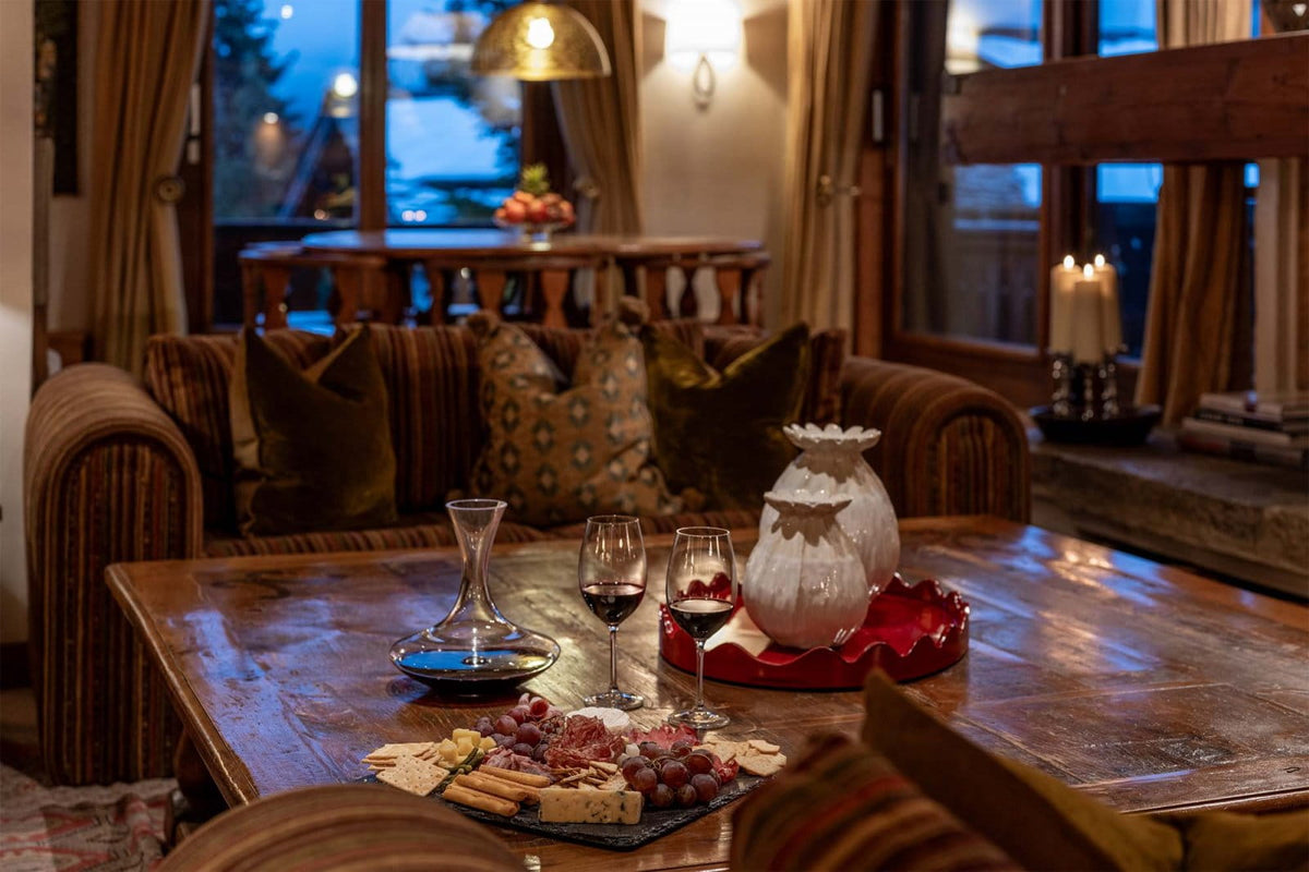 A cozy indoor dining or lounge scene with a wooden table set with wine glasses, a decanter, crackers, grapes, and a white decorative pitcher on a red tray. A striped upholstered sofa with patterned pillows sits behind the table, and warm wall lighting and large windows are visible in the background.
