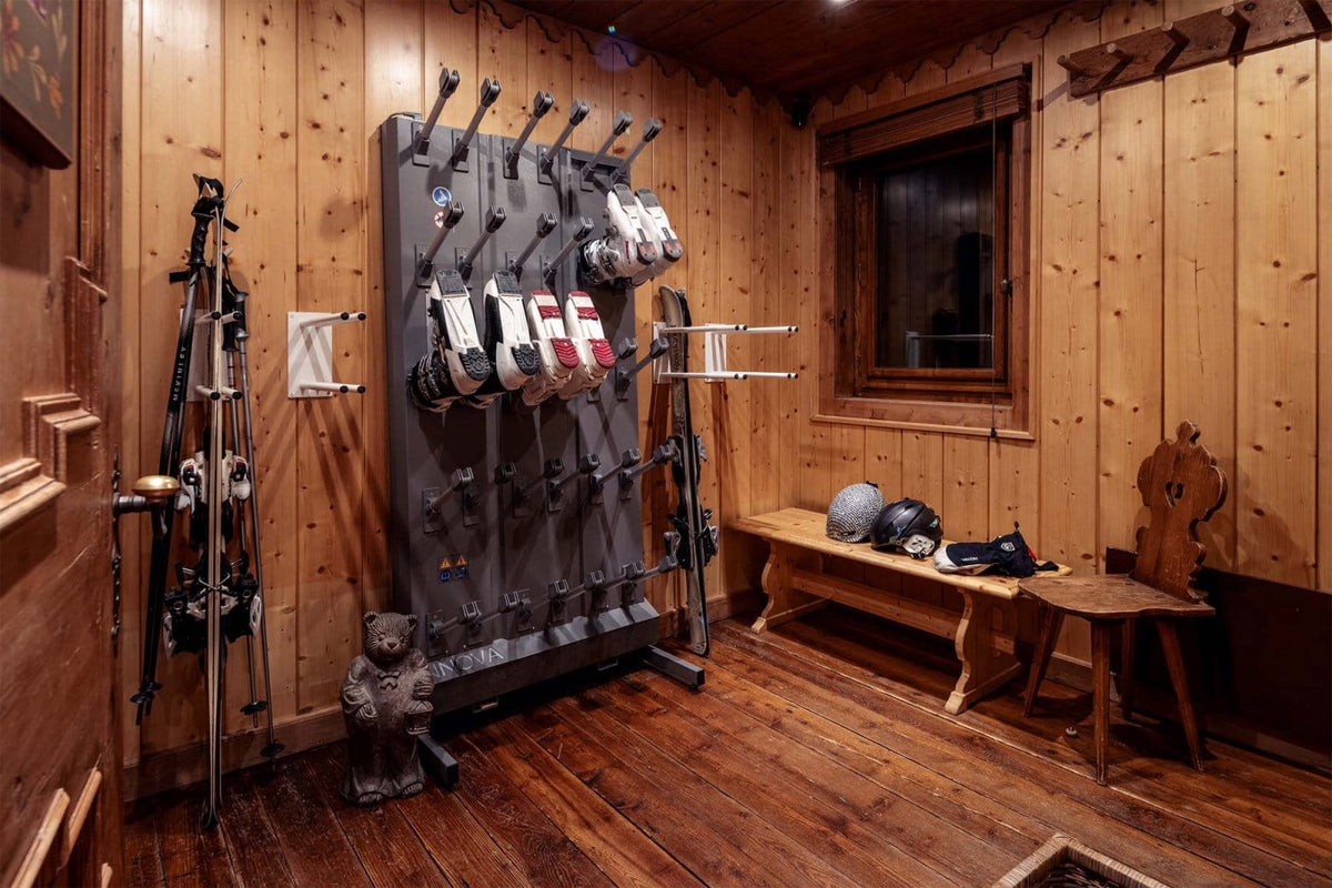 A wooden interior room with ski gear displayed on a large wall rack, including skis, boots, helmets, and poles. A wooden bench, stool, window, and wood-paneled walls are visible.