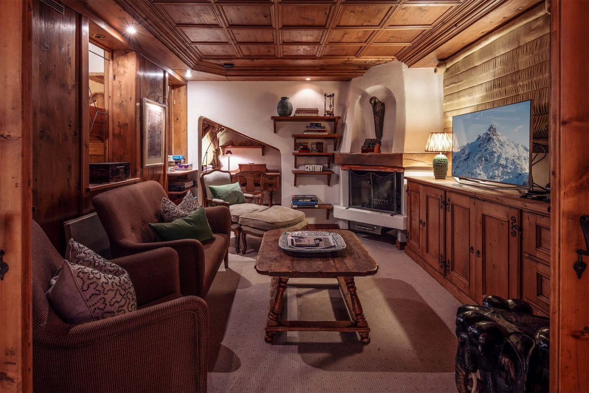 A cozy, rustic mountain chalet living room with warm wood paneling on the ceiling and walls, featuring brown upholstered armchairs, a wooden coffee table, a fireplace, and a television displaying a snowy mountain scene.