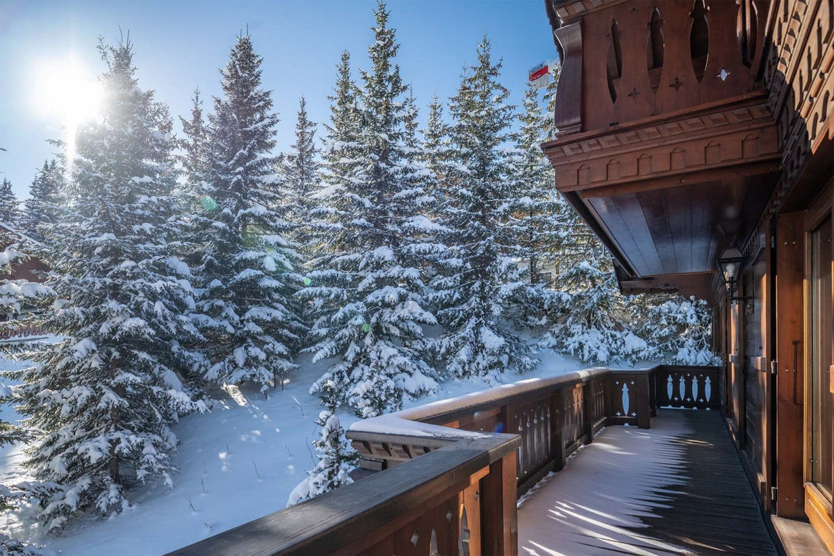 A snowy mountain chalet balcony with dark wood railings overlooking a snow-covered pine forest under a bright blue sky with the sun shining.