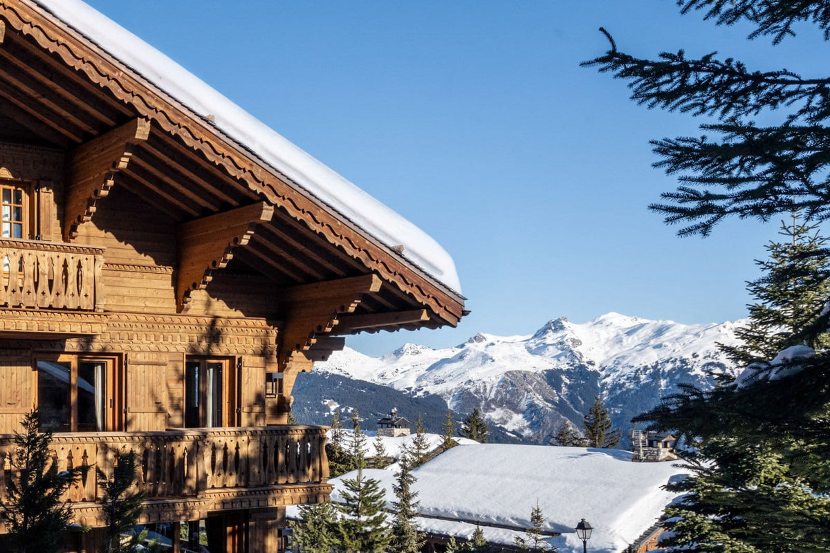 A traditional wooden chalet with snow-covered roofs in a mountain village, with snow-capped peaks and evergreen trees under a clear blue sky.