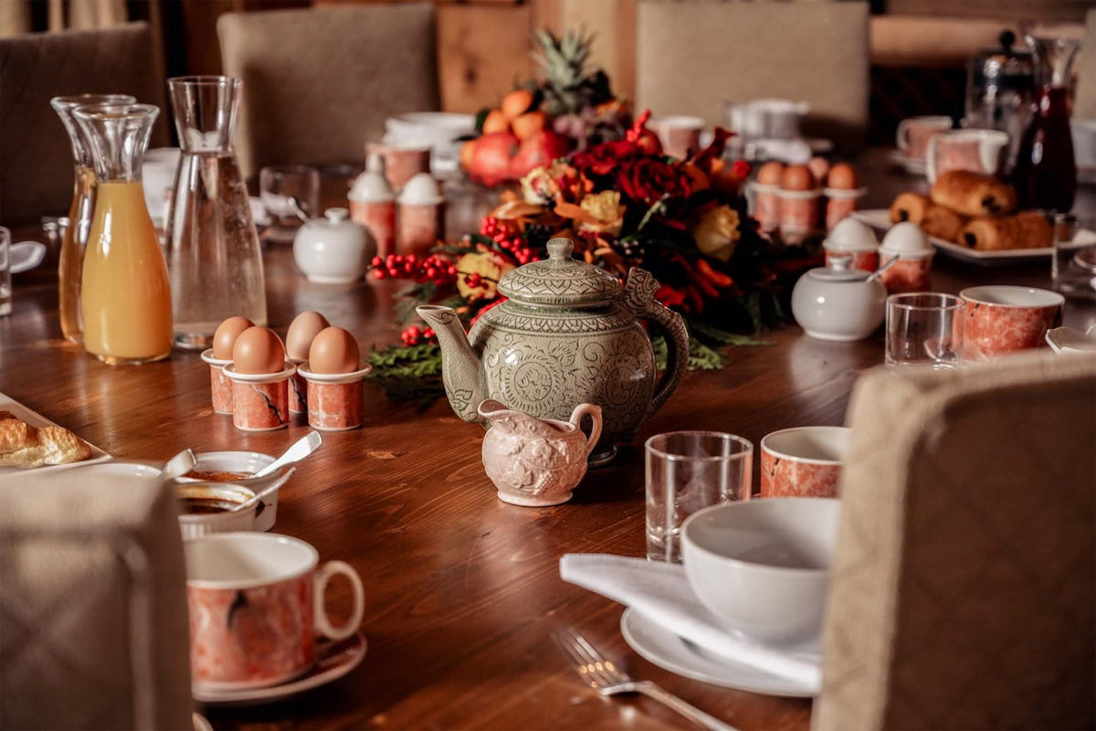 A styled breakfast table setting with a central patterned green teapot, white ceramic bowls and cups, copper-toned egg cups, glass carafes, and autumnal floral centerpieces on a wooden table.