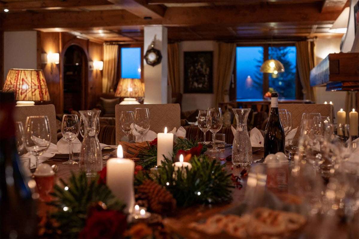A warmly lit dining table set for guests with lit white pillar candles, clear glass carafes and wine glasses, pine garlands, and red berries in a rustic indoor room with wooden ceiling beams and windows showing blue evening light outside.