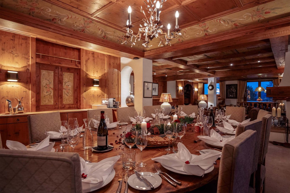 A warmly lit dining room with a long wooden table set for multiple guests, including wine glasses, folded white napkins, a bottle, and candles. The room features wood-paneled walls, a decorative chandelier, and cozy ambient lighting.