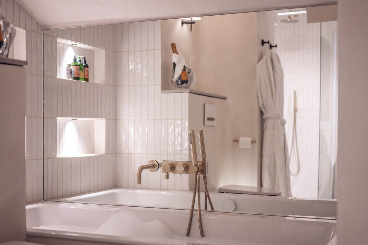 A modern white bathtub with a matte brass faucet set in a tiled bathroom, with a large mirror reflecting a white robe in a walk-in shower area.