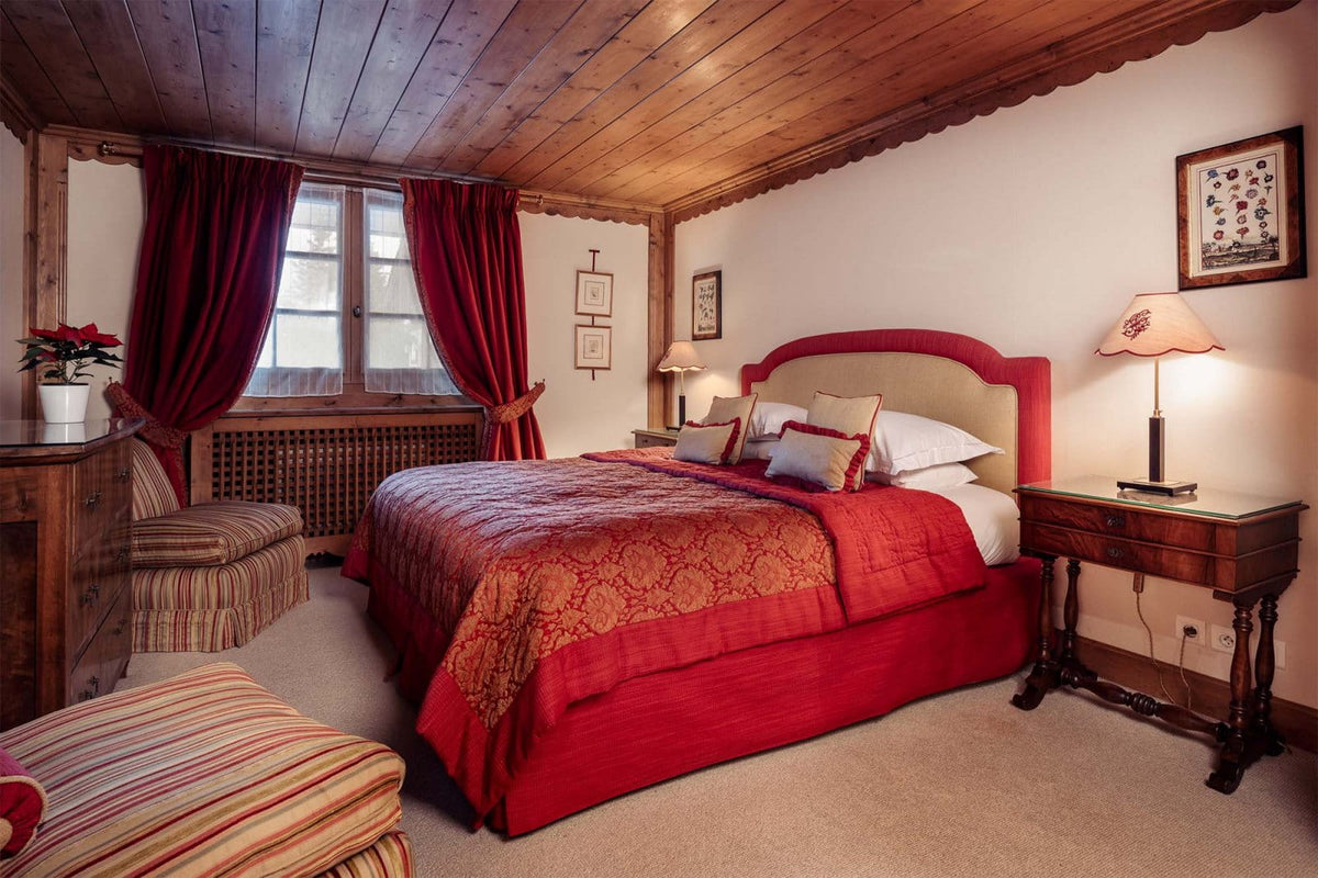 A cozy bedroom with a double bed dressed in red bedding and patterned covers, set in a rustic room with a wood-paneled ceiling, red curtains, bedside tables, and lamps.