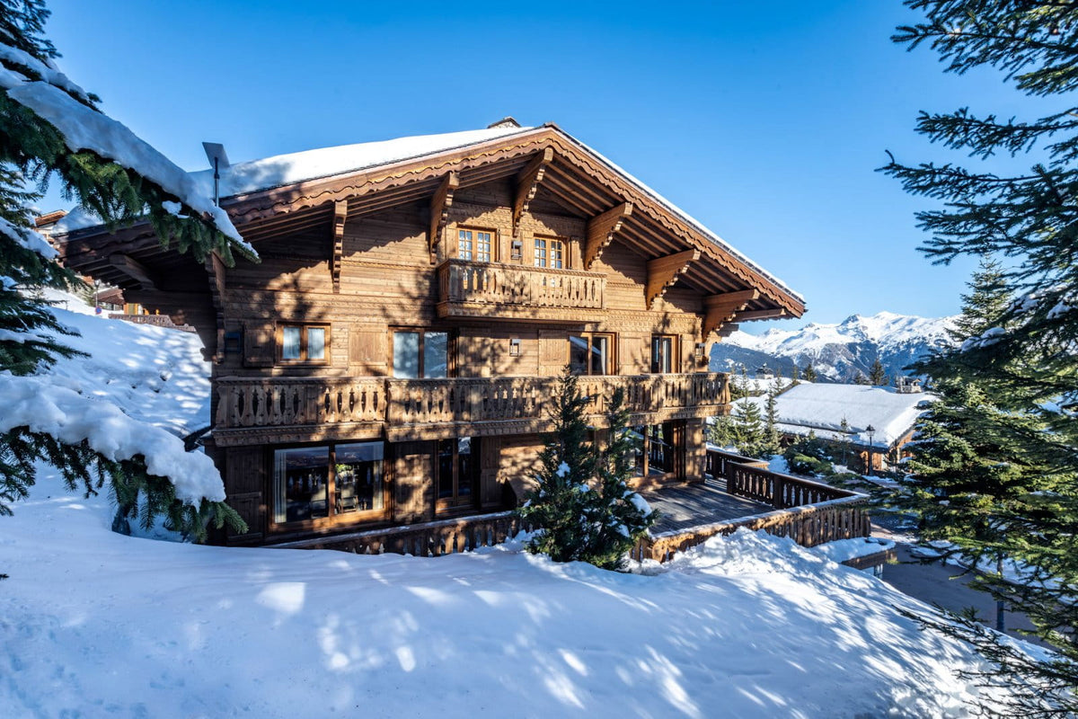 A large traditional alpine chalet with snow-covered roofs and balconies, surrounded by deep snow, evergreen trees, and distant mountains under a clear blue sky.