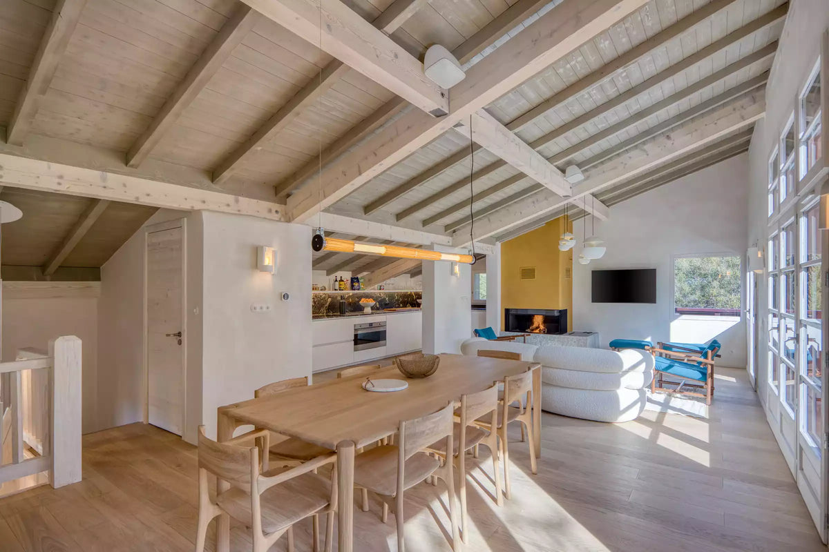 A spacious, rustic chalet-style living room with light wood walls and ceiling beams, featuring a large light gray sectional sofa, a wooden coffee table, a stone fireplace, and a starburst chandelier.