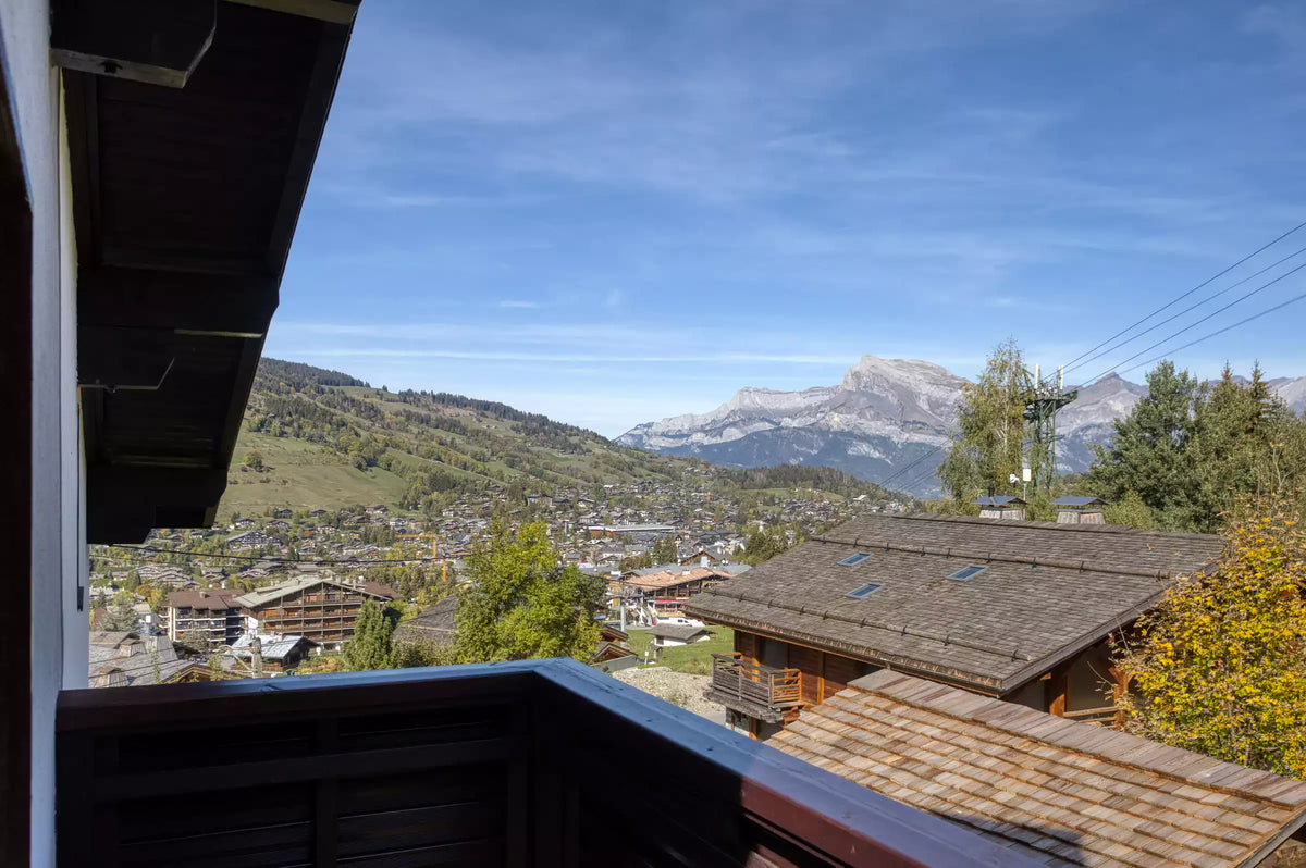 A scenic view from a balcony overlooking a mountain village with alpine chalets, green hills, and snow-capped mountains under a clear blue sky.