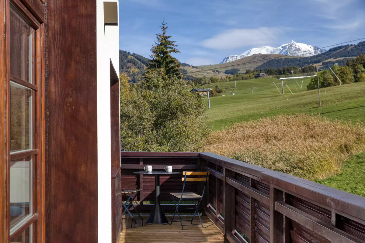 A wooden balcony overlooking a lush green mountain valley with snow-capped peaks under a blue sky, with a small table and chairs on the deck.