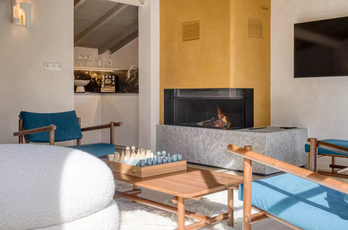 A modern living room interior featuring a black fireplace with a gray stone base, a mustard yellow wall, wooden chairs with blue cushions, a light wood coffee table, and a white textured sofa in the foreground.