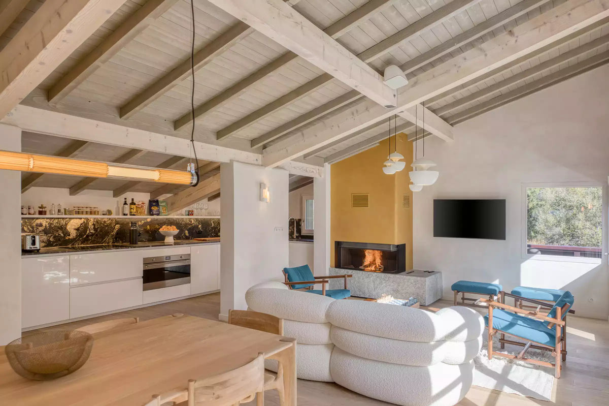 A bright, modern open-plan living space with exposed wooden ceiling beams, a white curved sofa, blue accent chairs, a built-in fireplace, and a kitchen area in the background.