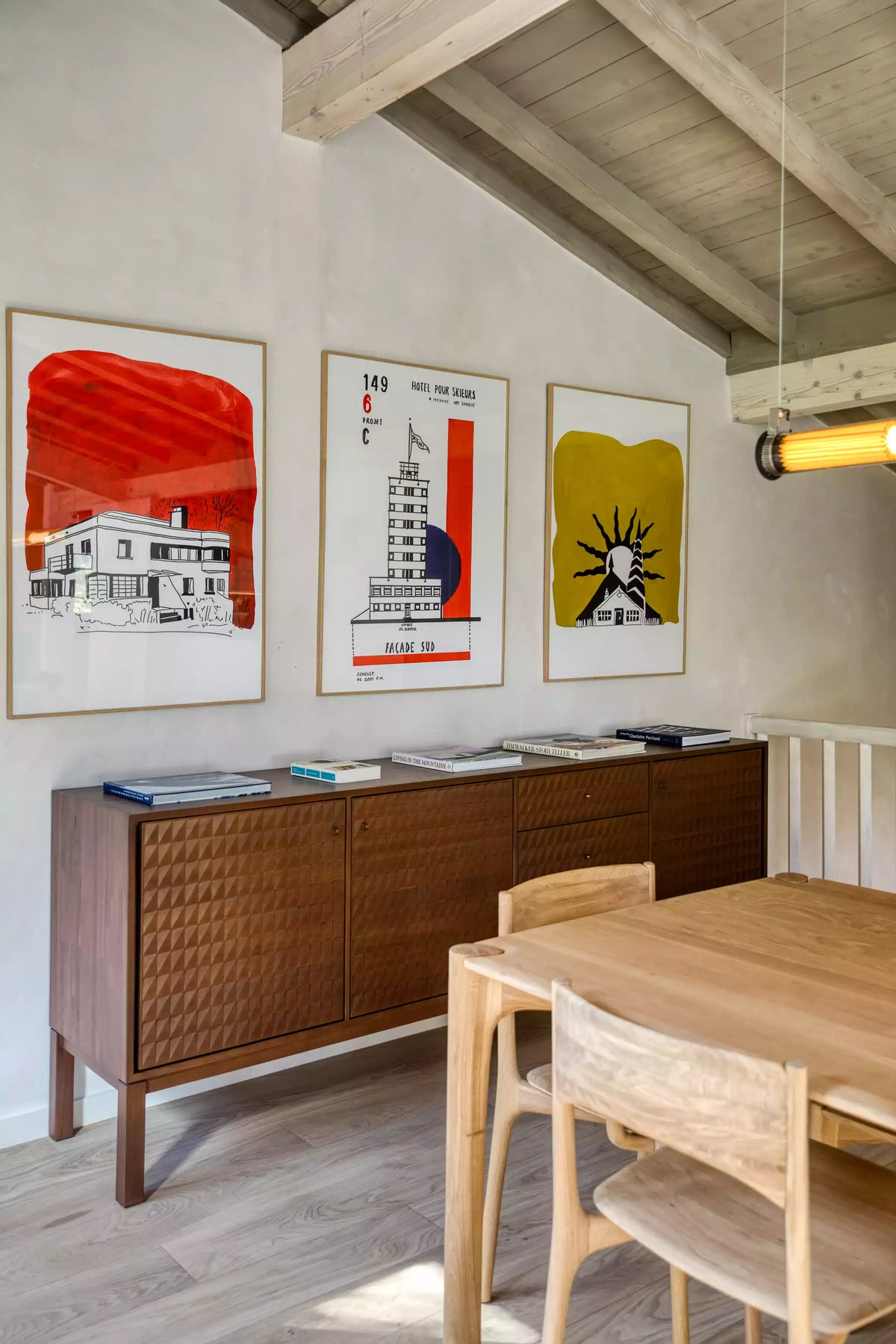 A styled interior room with three framed art prints on a white wall above a mid-century wooden sideboard, next to a light wood dining table and chairs under a slanted ceiling with exposed beams.