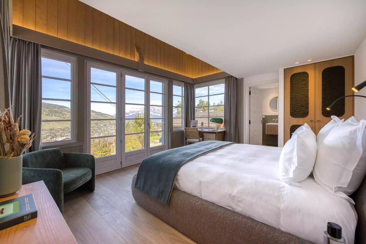 A modern bedroom interior featuring a large bed with white linens and a dark teal throw, set in a bright room with wood-paneled walls, floor-to-ceiling windows, a green armchair, and a view of outdoor hills.