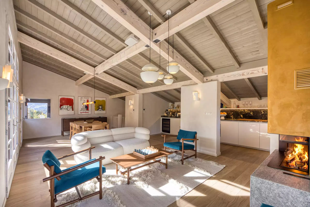 A bright, modern mountain-style living room with light gray sectional sofas, a wooden fireplace, and a large decorative sunburst chandelier hanging from exposed wooden beams.