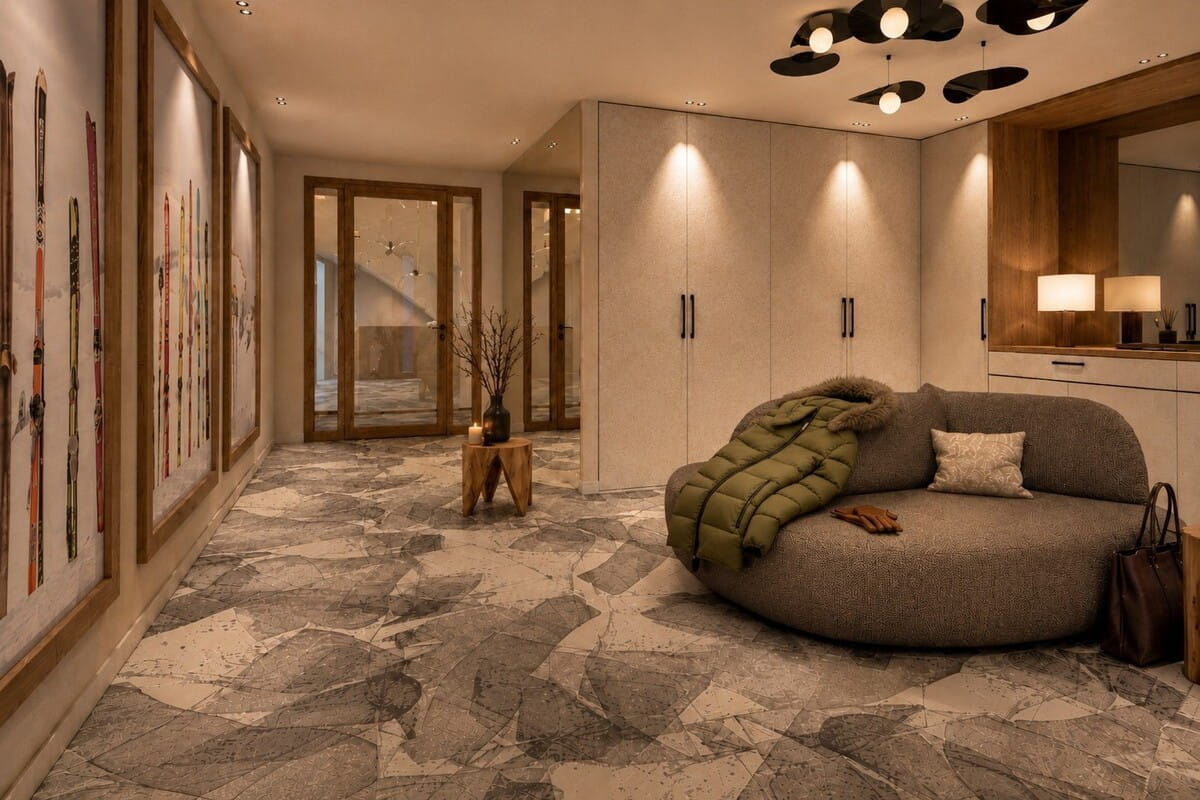 A modern interior space with a round upholstered lounge chair in the foreground, draped with an olive green quilted throw and a small pillow. The room has warm wood and light beige walls, a stone-patterned floor, wall-mounted lamps, and framed artwork along the left wall.