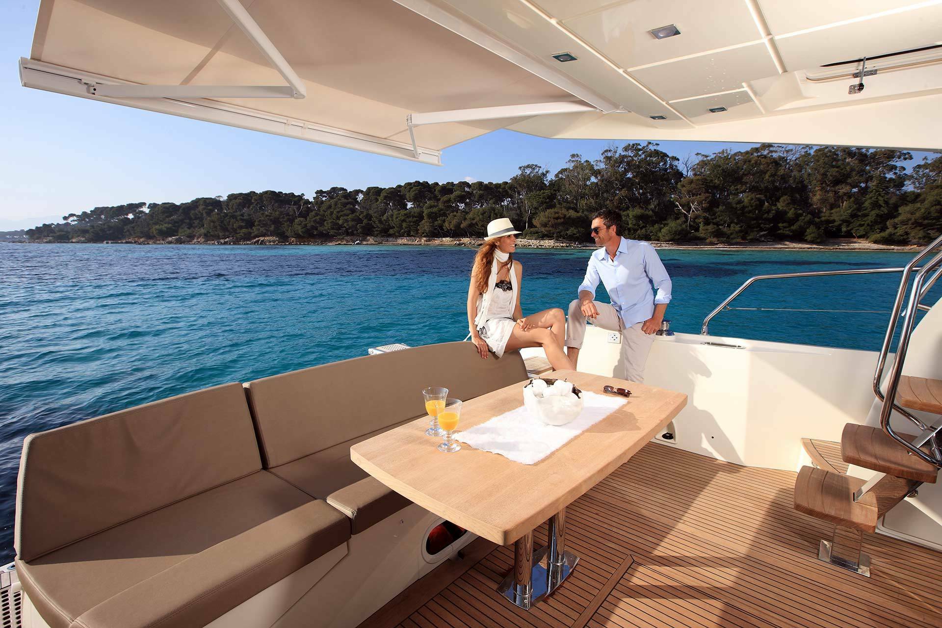 A man and a woman relax on the deck of a luxury yacht with a wooden table, beige seating, and a tropical ocean backdrop.