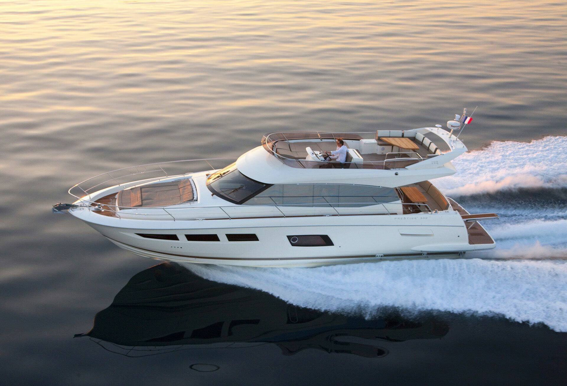 A luxury motor yacht with a white hull and warm wood accents speeds across dark water, creating a white wake. A person is visible on the upper deck.