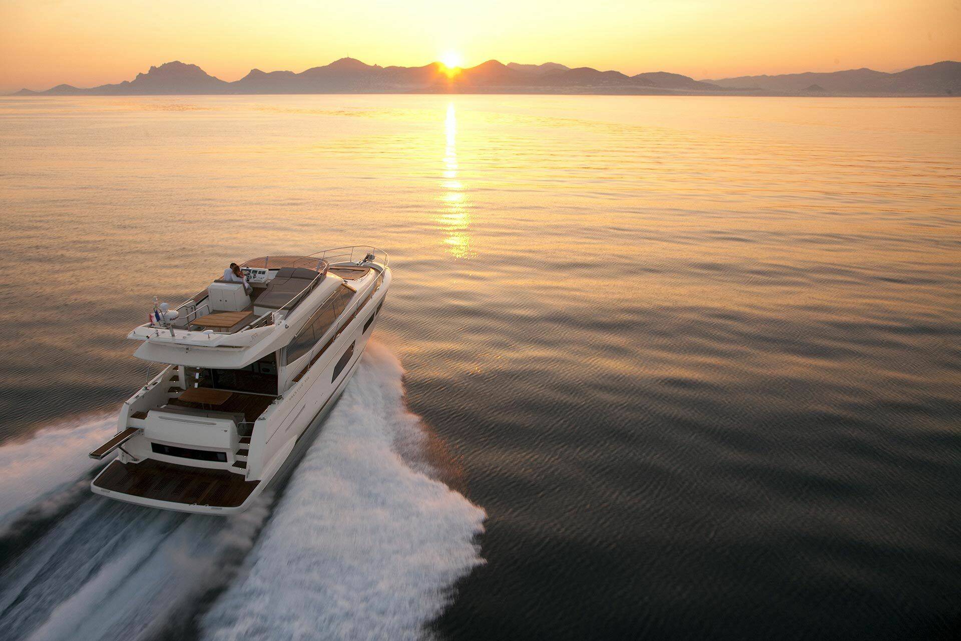 A luxury motor yacht speeds across calm ocean water at sunset, leaving a white wake behind it with distant mountains on the horizon.