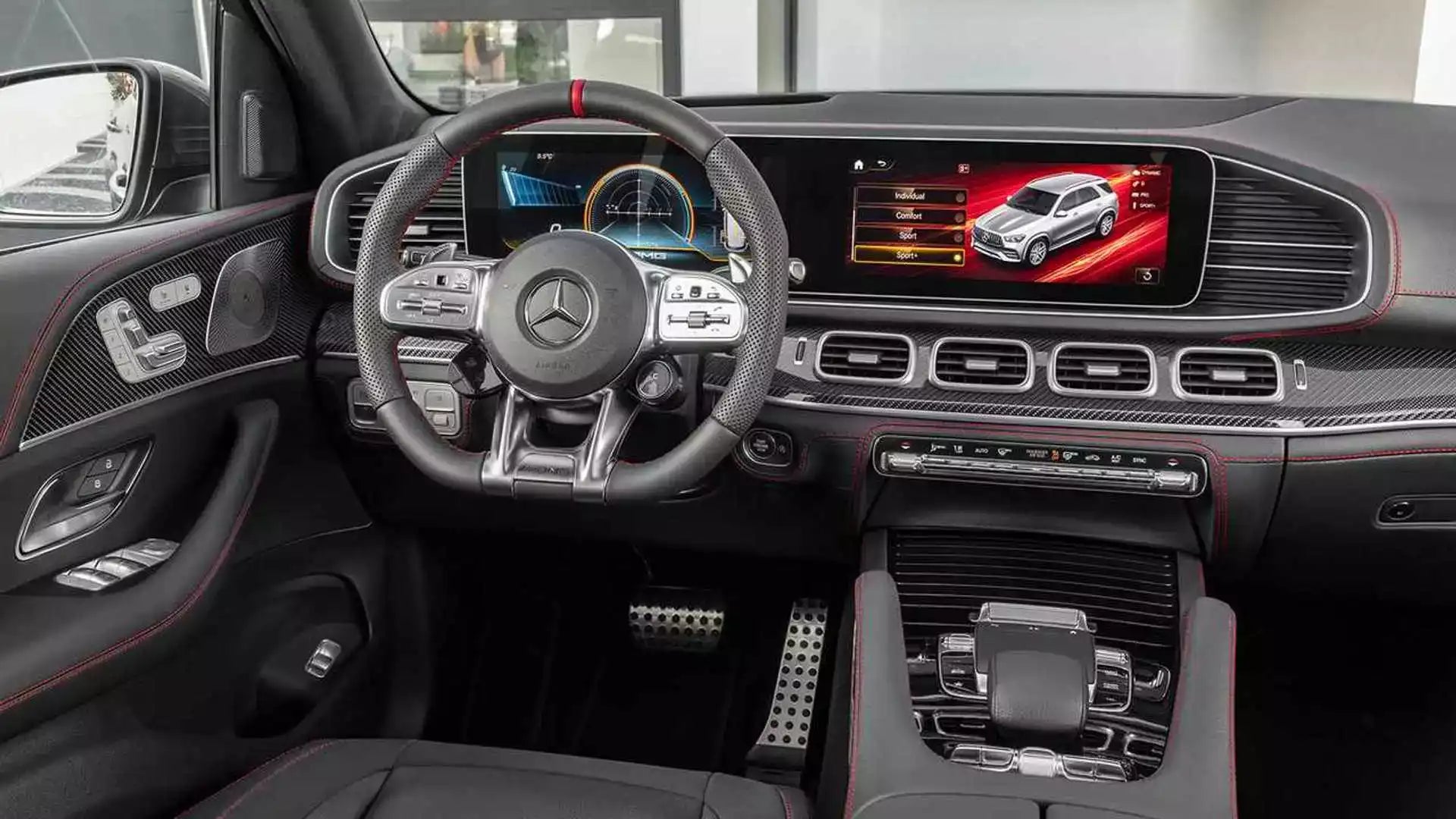 A luxury car interior featuring a black leather steering wheel with a silver Mercedes-Benz logo, a digital instrument cluster, a large infotainment screen, carbon fiber trim, and red stitching accents.
