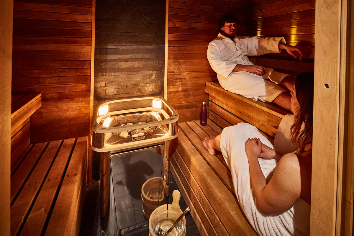 A traditional wooden sauna interior with two people wrapped in white robes sitting on wooden benches, with a metal steam basket and sauna accessories visible.