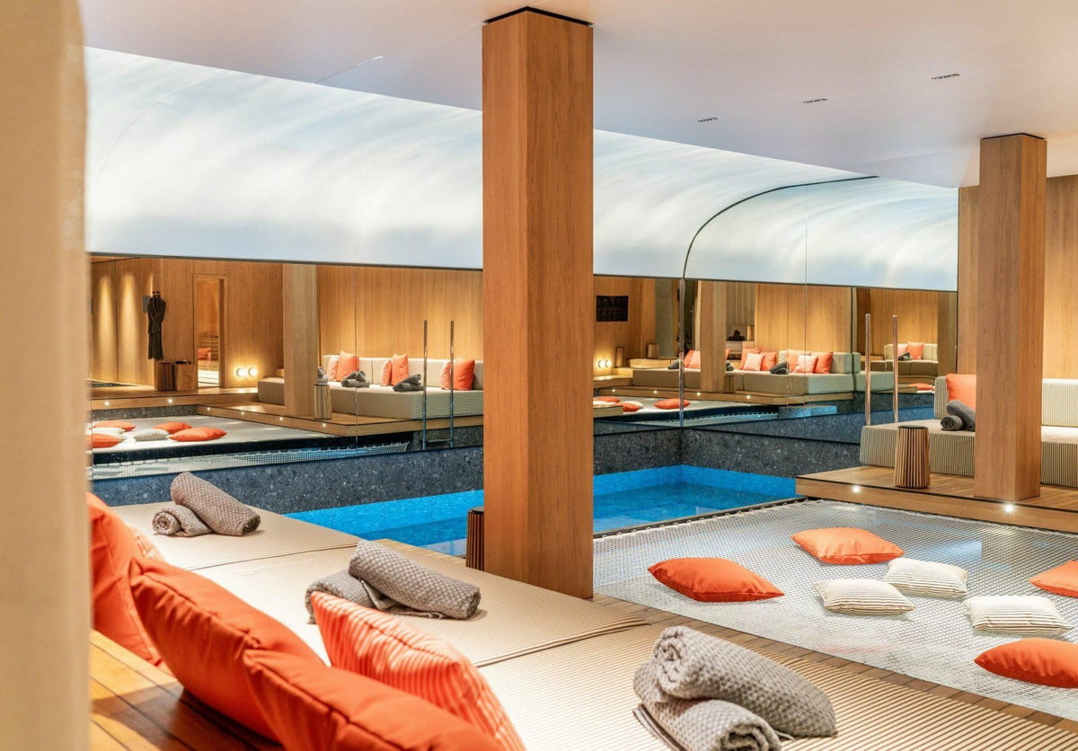 Luxury indoor poolside lounge area with wooden pillars, blue swimming pool, and orange and gray pillows on a light-colored deck.