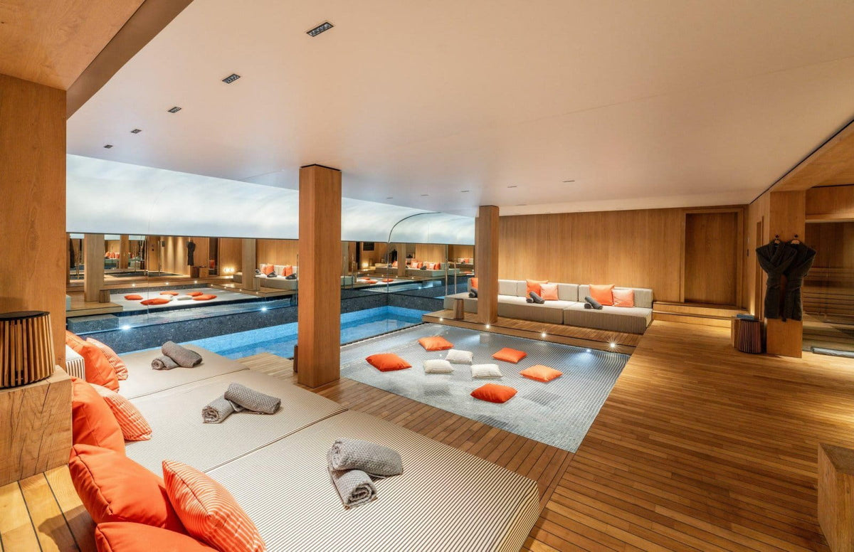 Luxury indoor poolside lounge area with a rectangular swimming pool, wooden columns, floor cushions, orange and white pillows, and a spa-like setting.