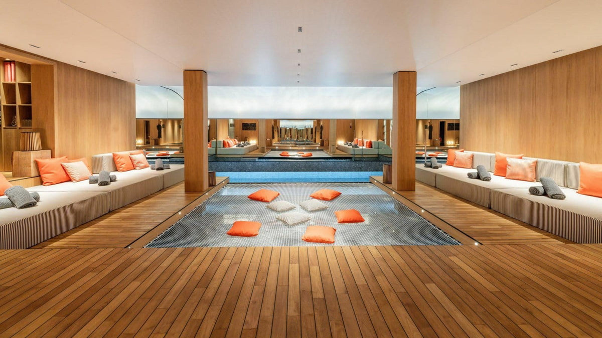 A luxury indoor pool area with a long rectangular pool, wooden deck flooring, built-in lounge seating with orange and gray pillows, and warm wood paneling.