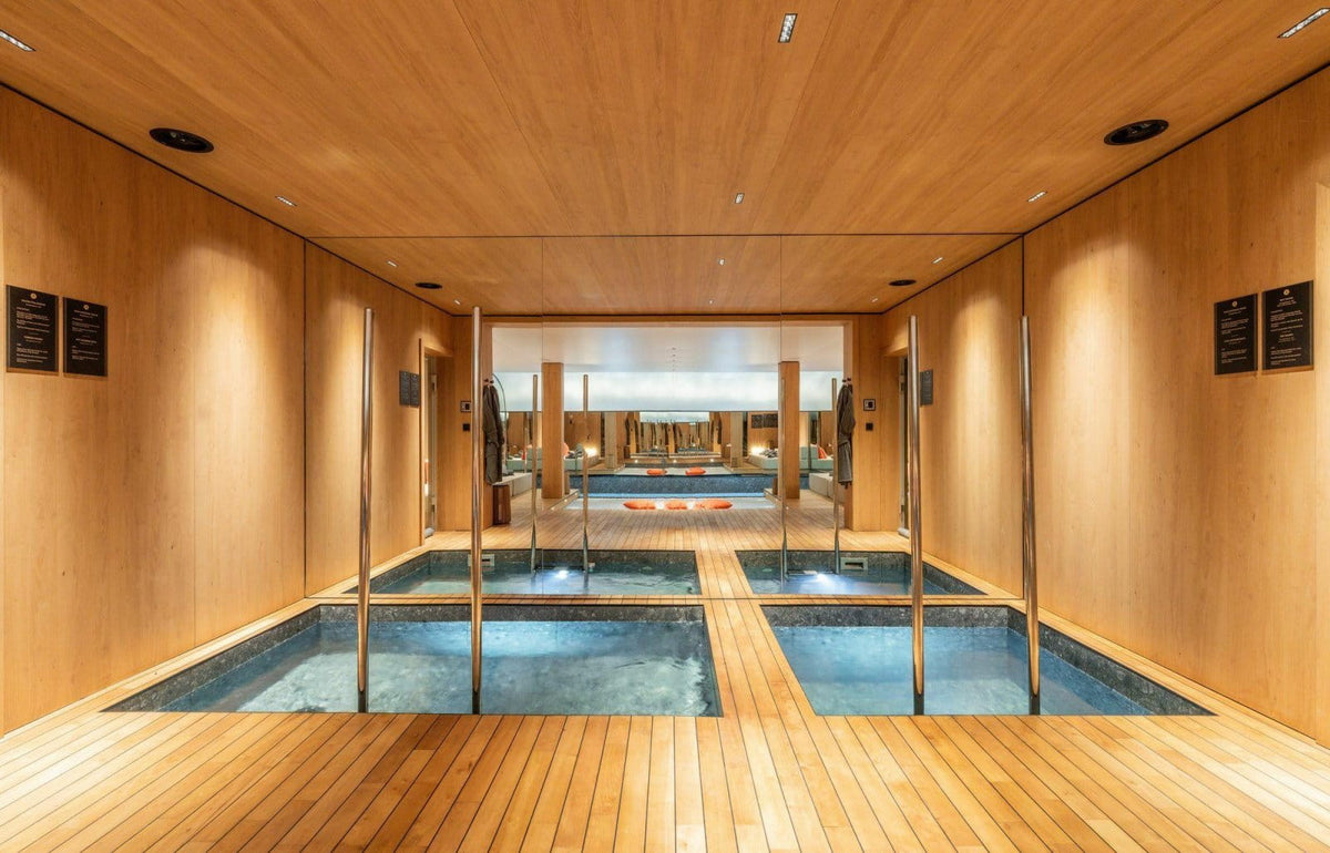 An indoor spa or pool area with light wood walls and ceiling, glass dividers, and rectangular blue water pools on a wood-look floor.
