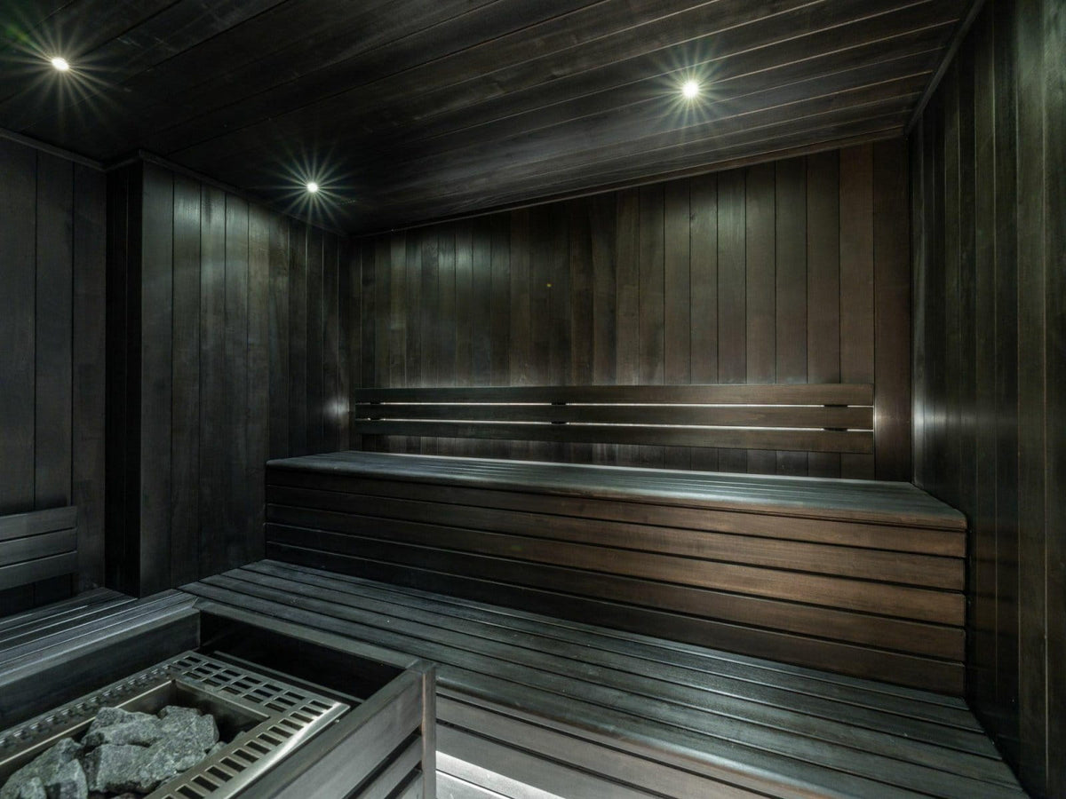 Dark wood sauna interior with tiered wooden benches, wall panels, ceiling lights, and a stone firebox in the foreground.