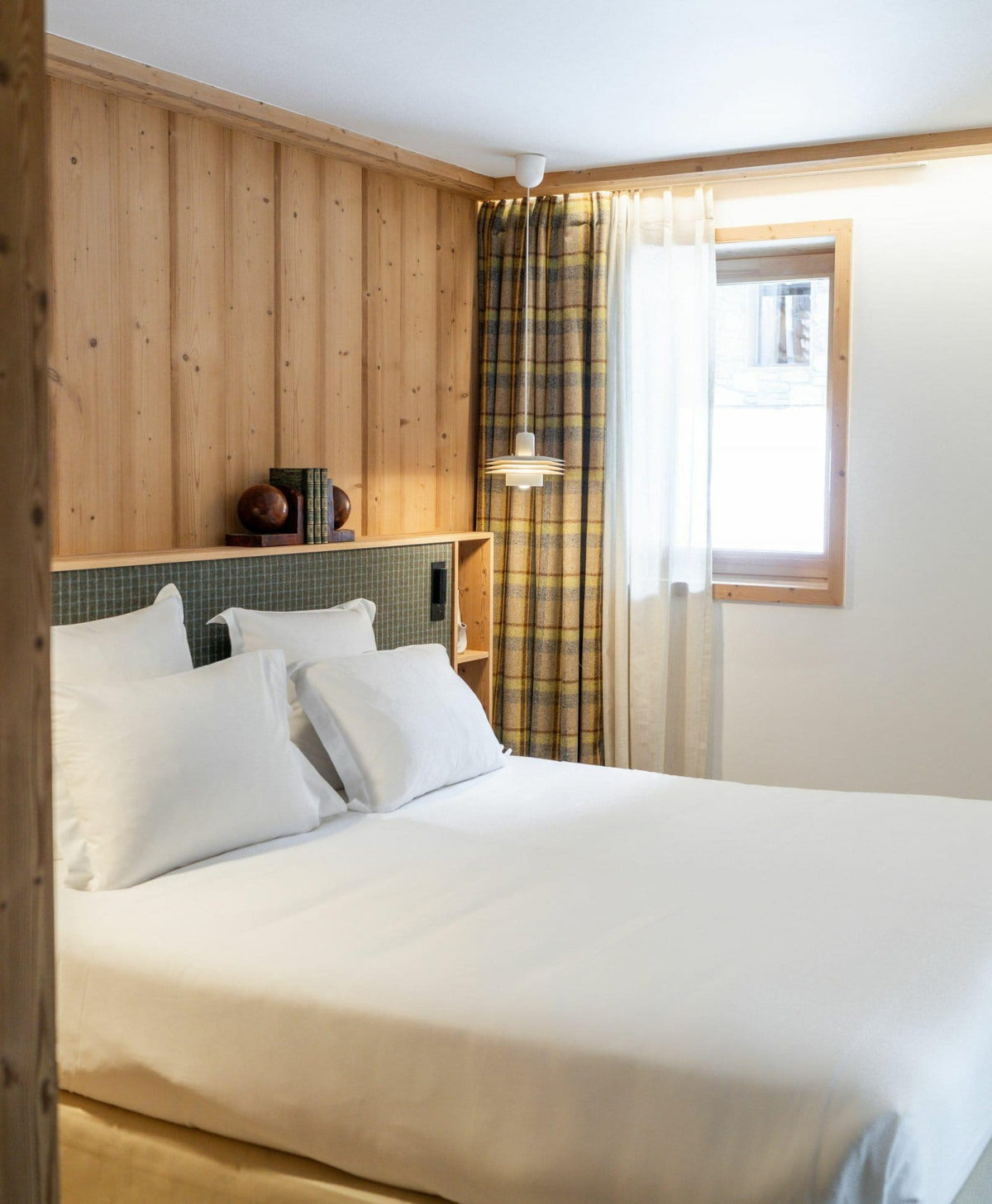A cozy bedroom interior featuring a neatly made bed with white bedding, a light wood-paneled wall, a window with sheer white curtains and a yellow plaid accent curtain, and a wall-mounted lamp.