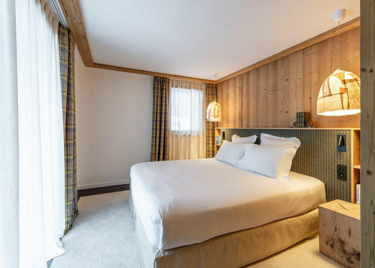 A modern bedroom interior featuring a neatly made bed with white linens, a wood-paneled headboard wall, hanging pendant lights, and large windows with sheer curtains and plaid drapes.