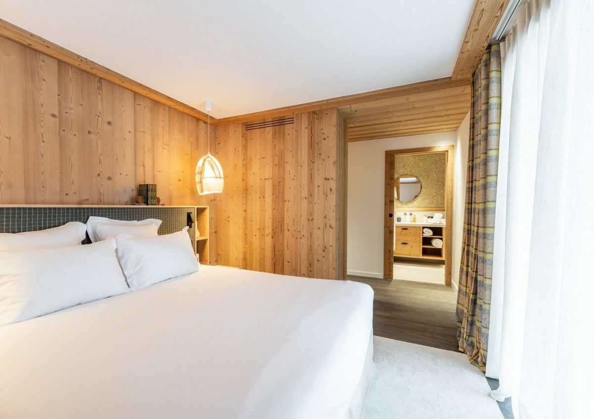 A modern bedroom with a large bed dressed in white linens, set against warm wood-paneled walls. A hanging pendant light illuminates the bed area, and an open doorway in the background reveals a bathroom.