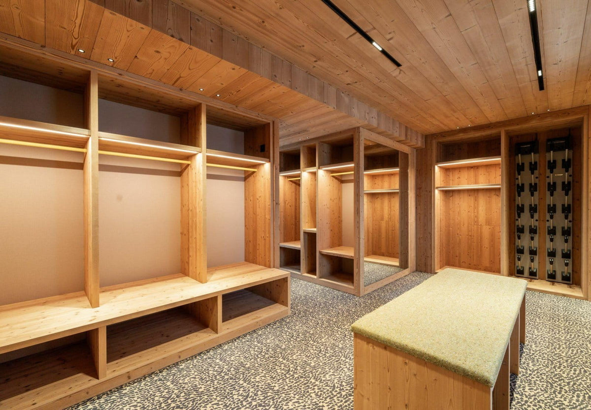 A luxury walk-in closet room featuring custom wooden shelving, a matching wood ceiling, a textured gray and white speckled floor, and a central wooden vanity table with a green upholstered top.