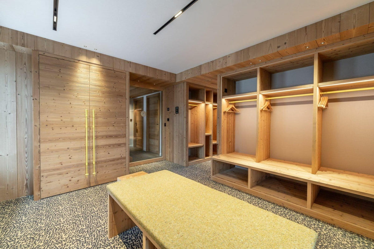 A modern wooden walk-in closet with built-in shelving, hanging rods, bench seating, and a textured yellow-green bench in the foreground, set in a bright contemporary room with wood-paneled walls.