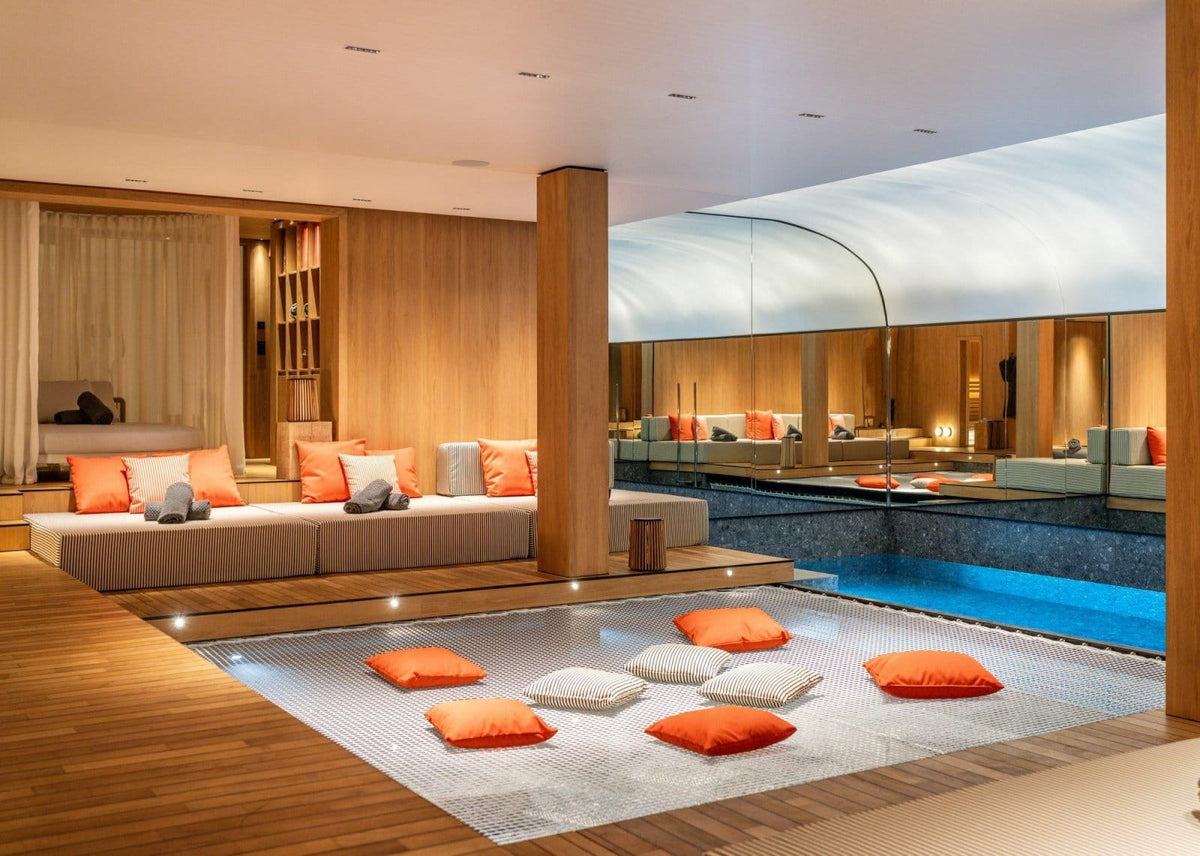 Luxury indoor resort pavilion with a modern wooden lounge set, orange and white pillows, a low blue swimming pool, and a light-colored floor mat with several orange and white floor cushions in the foreground.