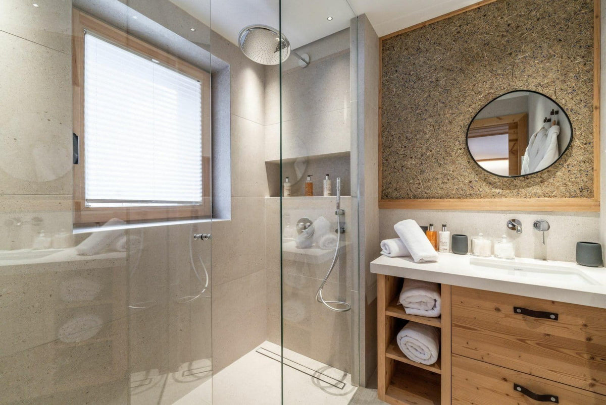 A modern bathroom interior featuring a glass shower enclosure, light wood vanity, white countertop, round mirror, and beige stone wall accents.