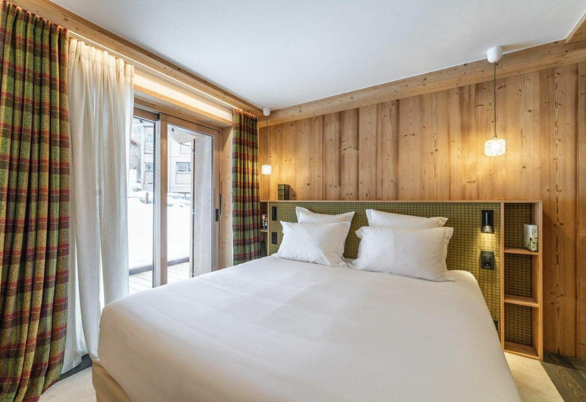 A cozy bedroom interior with a neatly made bed, white bedding, patterned green and red curtains, and a wood-paneled wall with built-in shelving.