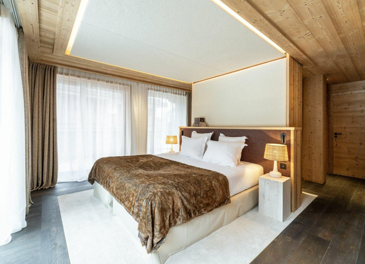 A modern mountain-style bedroom with a large bed dressed in white linens and a brown patterned throw blanket, set in a wood-paneled room with bedside tables, lamps, sheer curtains, and a white rug.