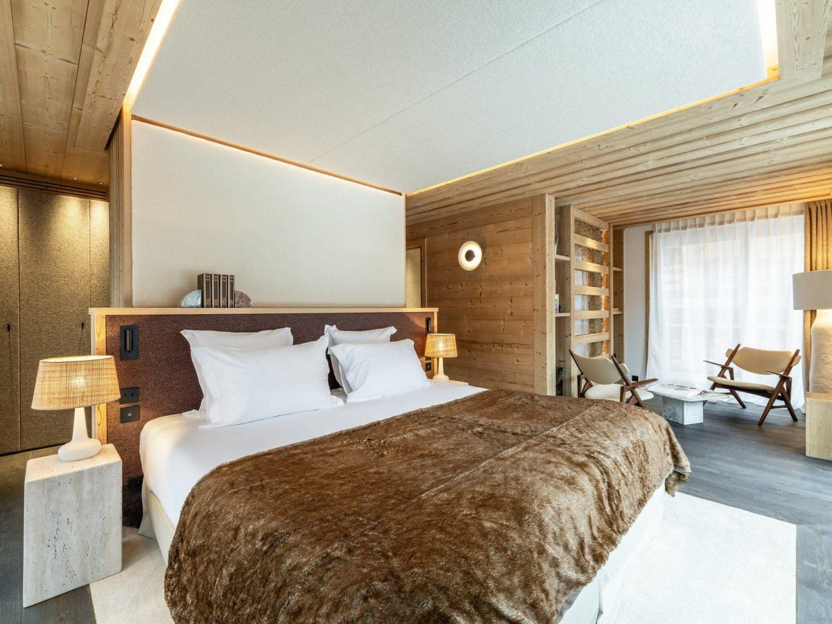 A modern mountain-style bedroom featuring a large bed with white linens and a brown faux-fur throw, set in a wood-paneled room with bedside tables, lamps, and a seating area by a window.