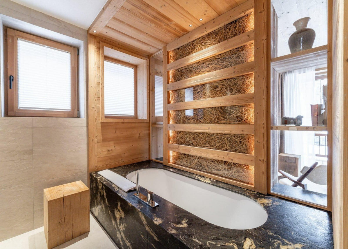 A luxury modern bathroom featuring a white oval bathtub set into a black and gold marble countertop, surrounded by light wood paneling and a decorative wall of woven straw baskets with warm lighting.