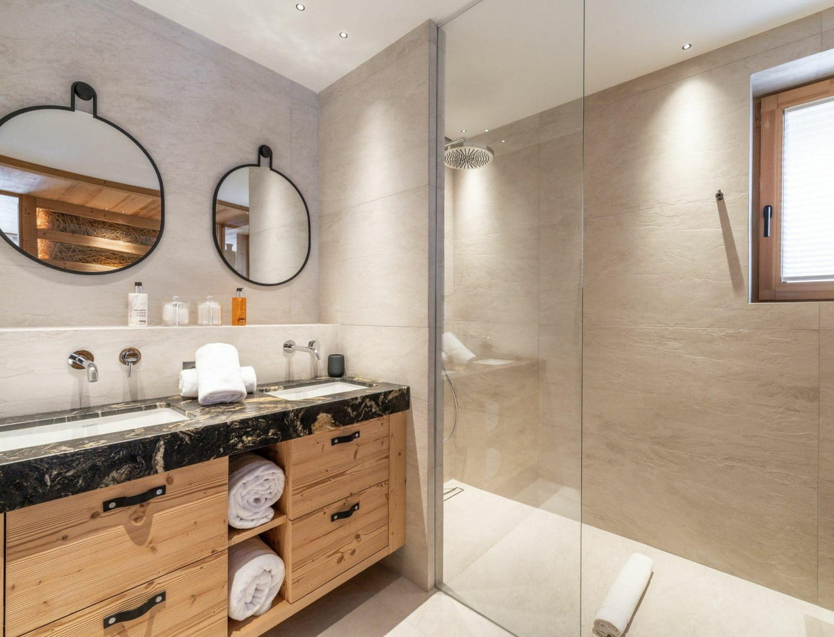 Modern bathroom interior with a wooden double vanity, black marble countertop, two round wall mirrors, a walk-in glass shower enclosure, and beige stone tile walls and floor.