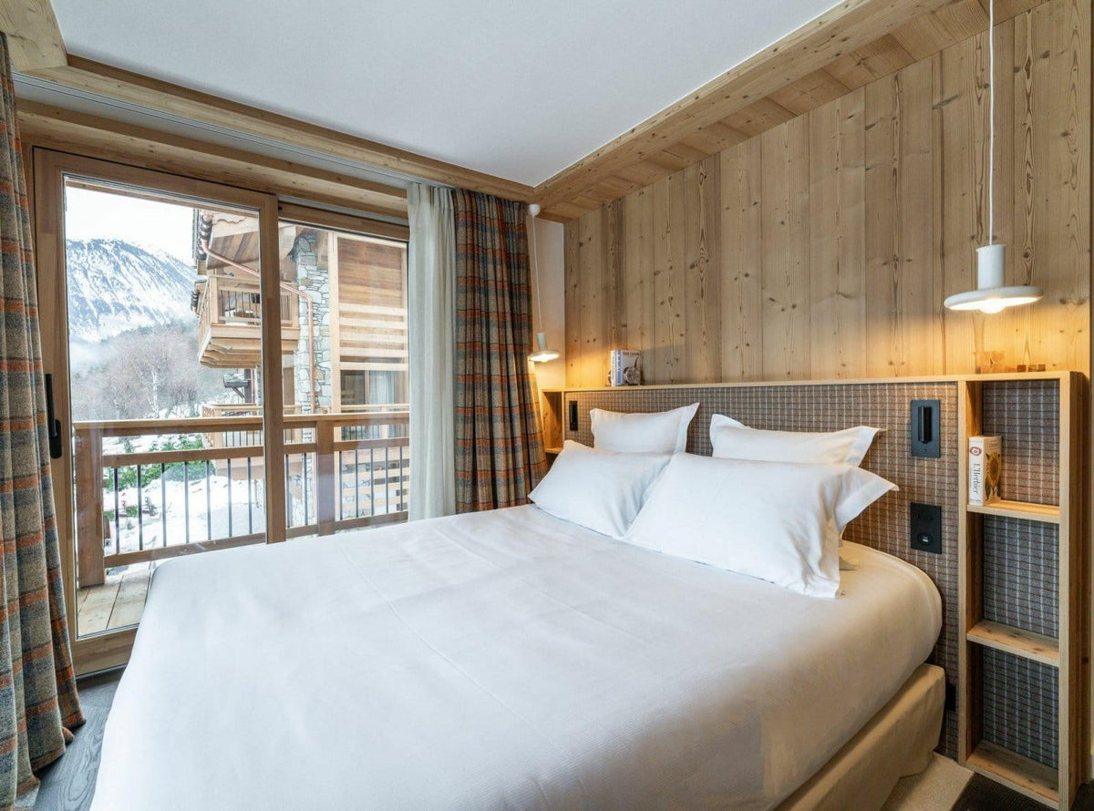 A modern ski chalet bedroom featuring a large bed with crisp white linens, set against a light wood-paneled wall with built-in shelving. A large sliding glass door on the left offers a view of a snowy mountain landscape.